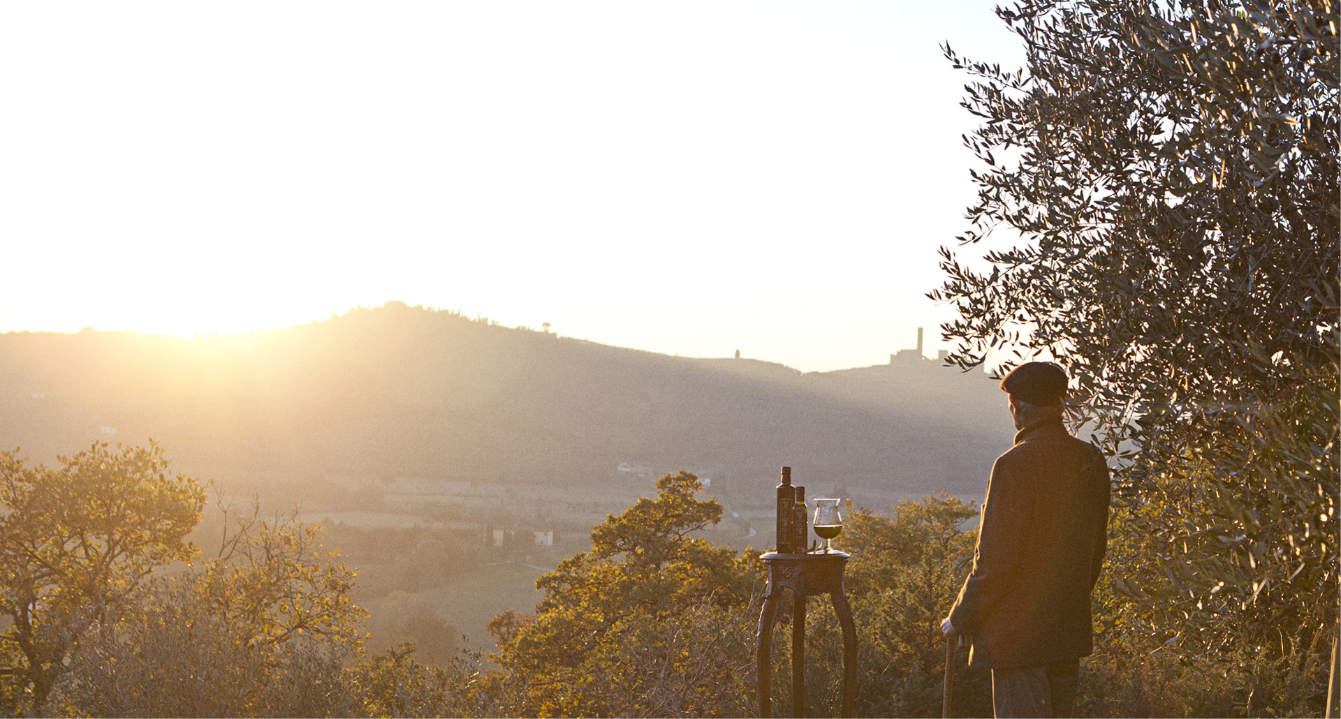 Uomo di spalle con cappello e cappotto che contempla un panorama collinare toscano al tramonto, accanto a un tavolino con una bottiglia e un calice di olio extravergine di oliva, con un ulivo in primo piano e torri medievali sullo sfondo avvolte nella luce dorata.