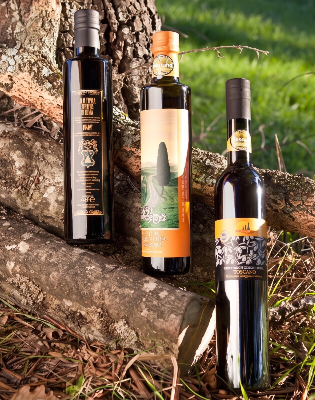 Tre bottiglie di olio extravergine di oliva Toscano IGP appoggiate a tronchi d'albero in un ambiente naturale: una con etichetta nera e dorata, una con etichetta arancione raffigurante un paesaggio toscano con cipressi, e una con etichetta nera e banda arancione con marchio IGP.