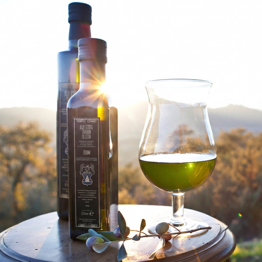 Bottiglia di olio extravergine di oliva con un calice contenente olio verde dorato, posati su una superficie in legno con un ramoscello di ulivo, al tramonto con paesaggio collinare sullo sfondo.