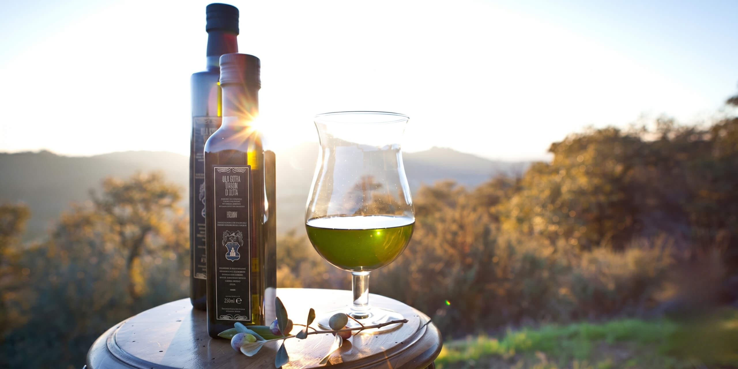 A bottle of extra virgin olive oil with a glass containing golden green oil, placed on a wooden surface with an olive branch, at sunset with a hilly landscape in the background.